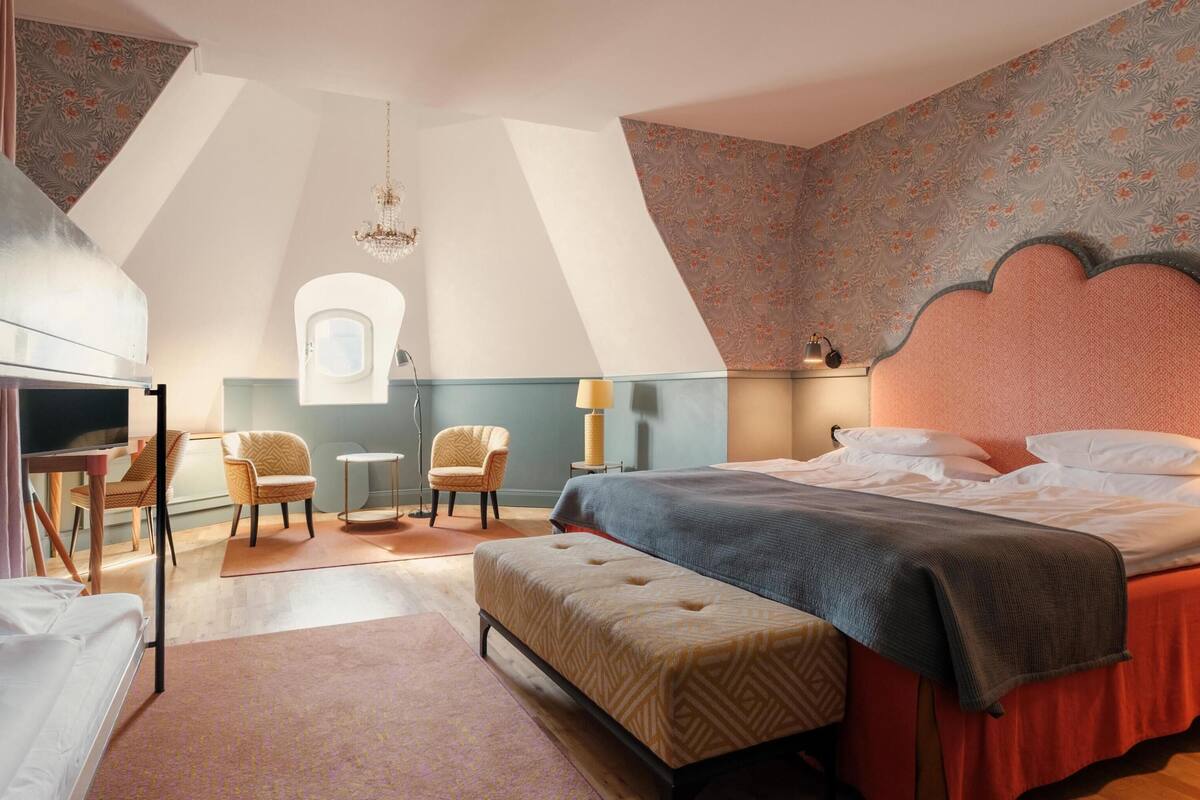 A bedroom with a bed, two chairs, a desk, a small table, a window, and a chandelier.