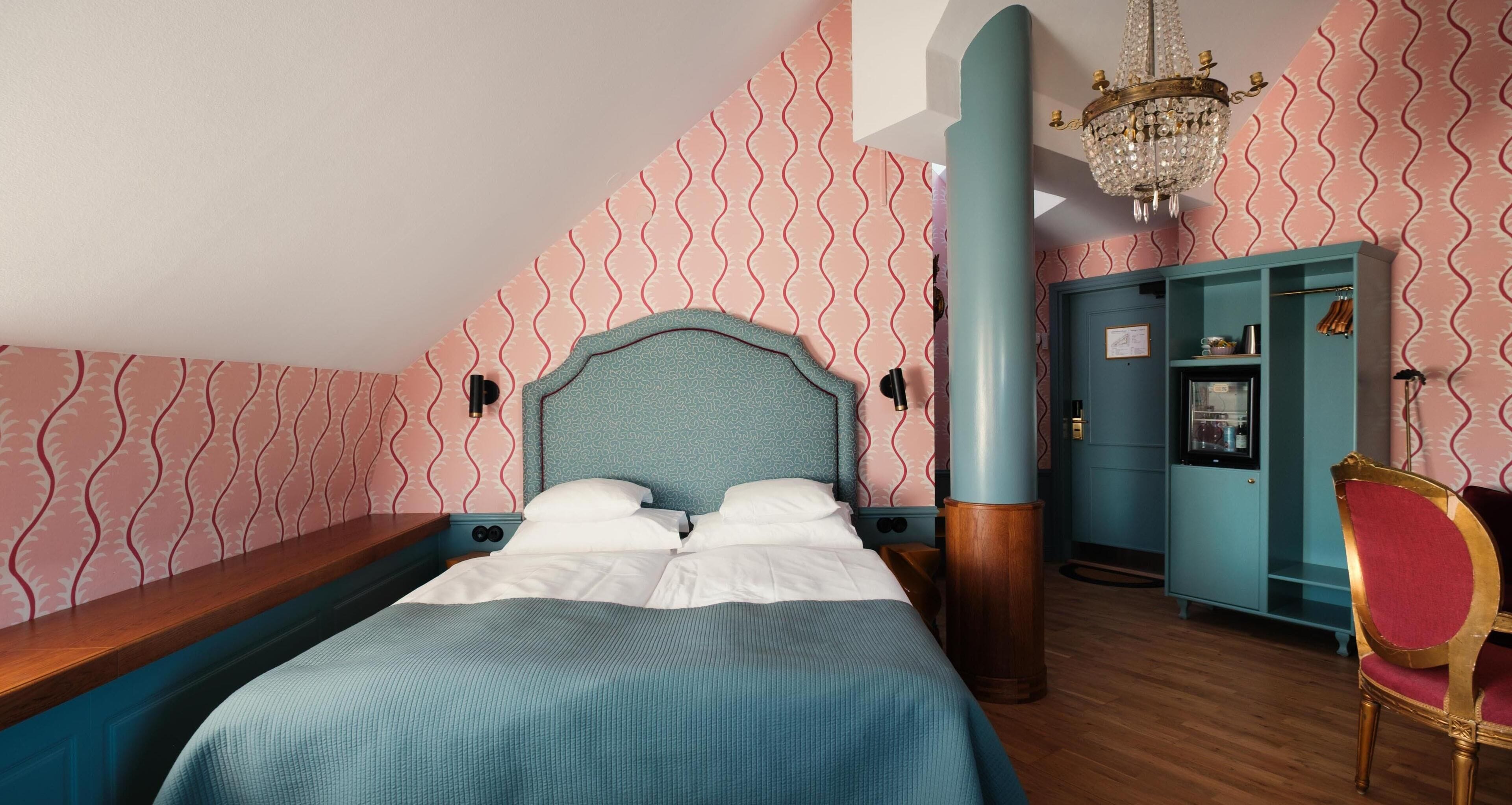 A bedroom with a bed, a wooden bench, a teal wardrobe, a red chair, and a chandelier.