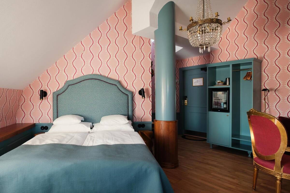 A bedroom with a bed, a wooden bench, a teal wardrobe, a red chair, and a chandelier.
