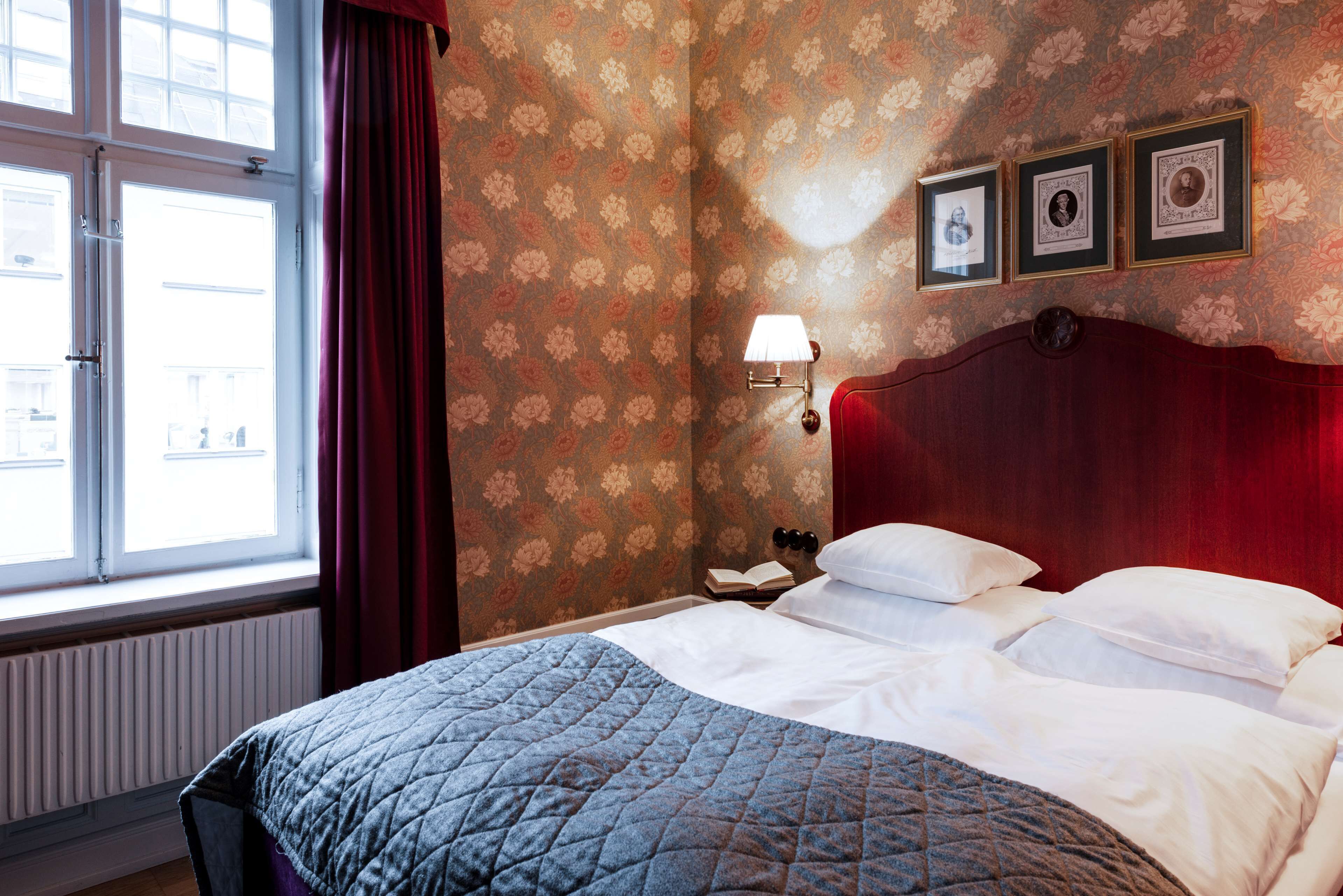 Una habitación de hotel con una cama, una ventana con cortinas, un radiador y tres cuadros en la pared.