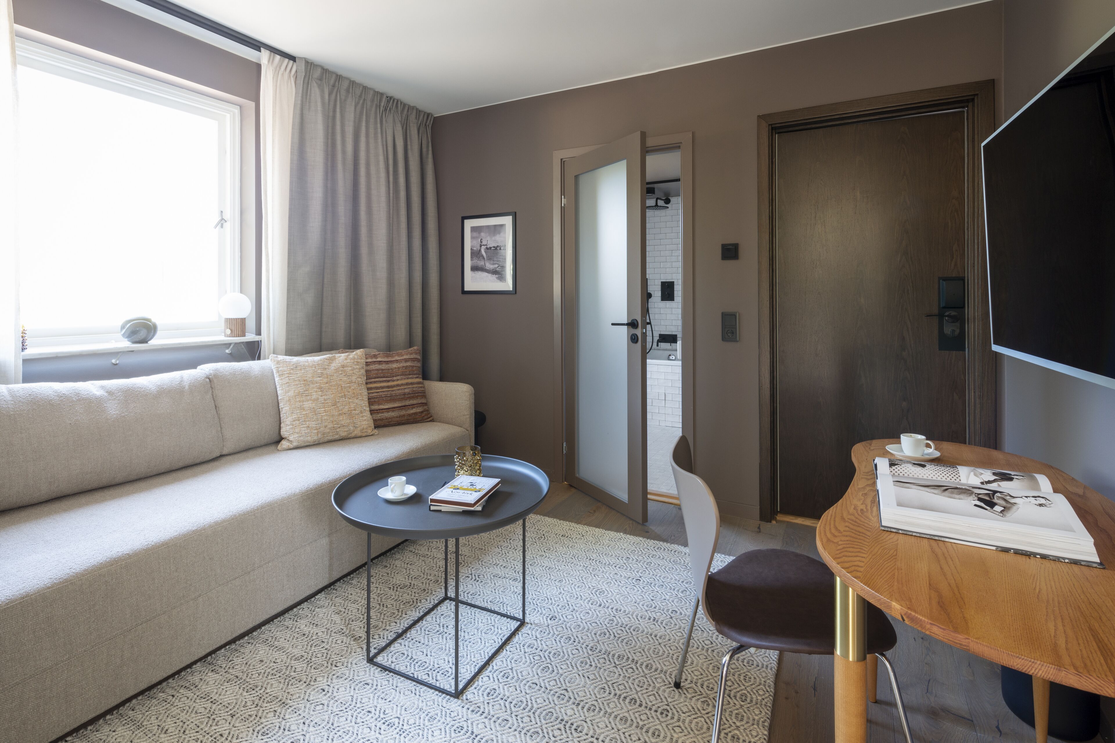 Ett modernt hotellrum med en soffa, ett litet runt bord, en stol, ett stort fönster med gardiner och en dörr som leder till badrummet.