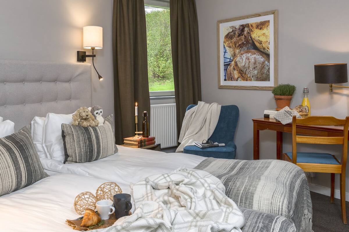 A bedroom with a large bed, a bedside table with books and a lamp, a chair, a desk with a lamp, and a window with curtains.