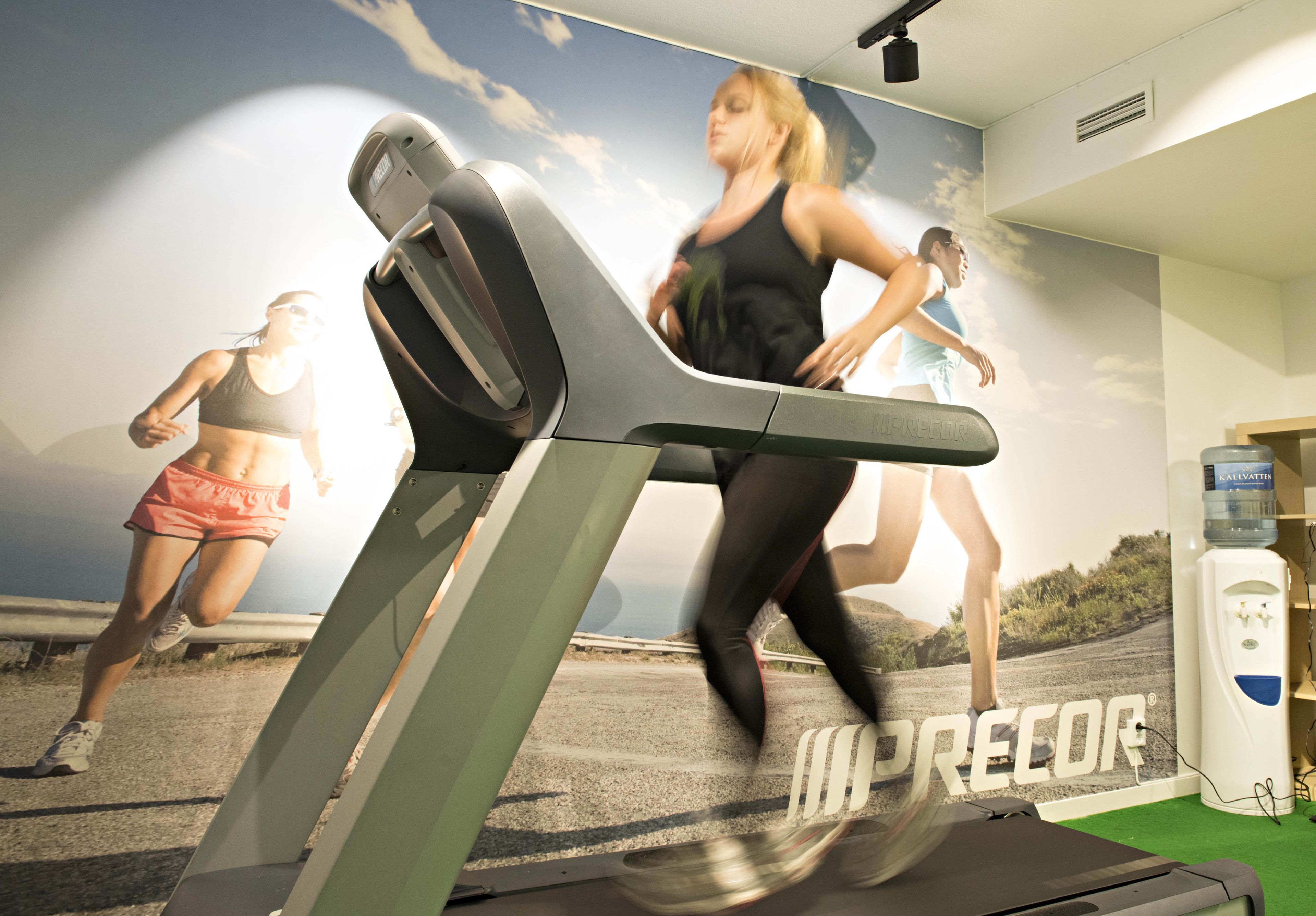 A treadmill with a 'PRECOR' logo, a water dispenser, and a wall mural of runners.
