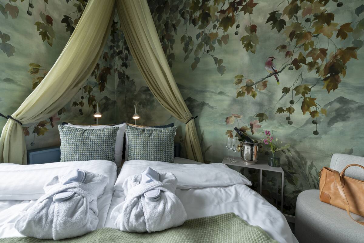 A hotel room with a bed, two pillows, a bedside table with a vase and a lamp, and a chair.