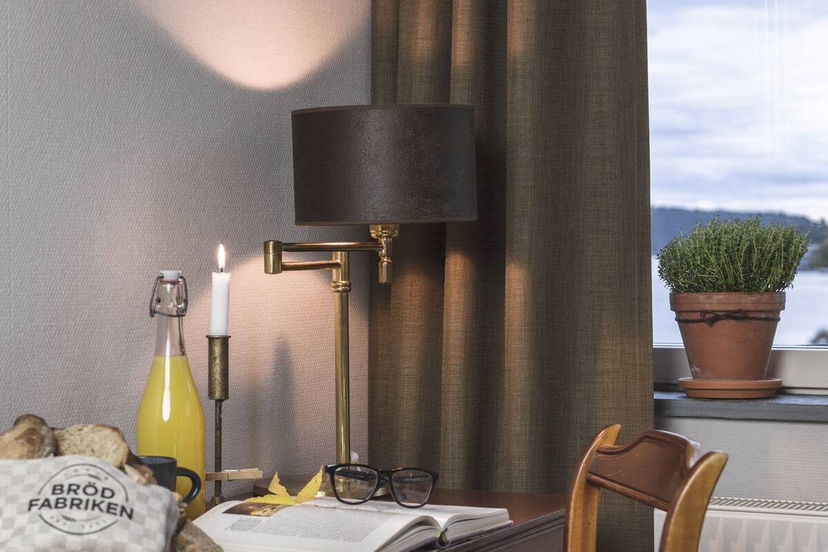 A wooden desk with a lamp, a candle, a bottle, and a bag labeled "Brod Fabriken" in a room with a view of the water.