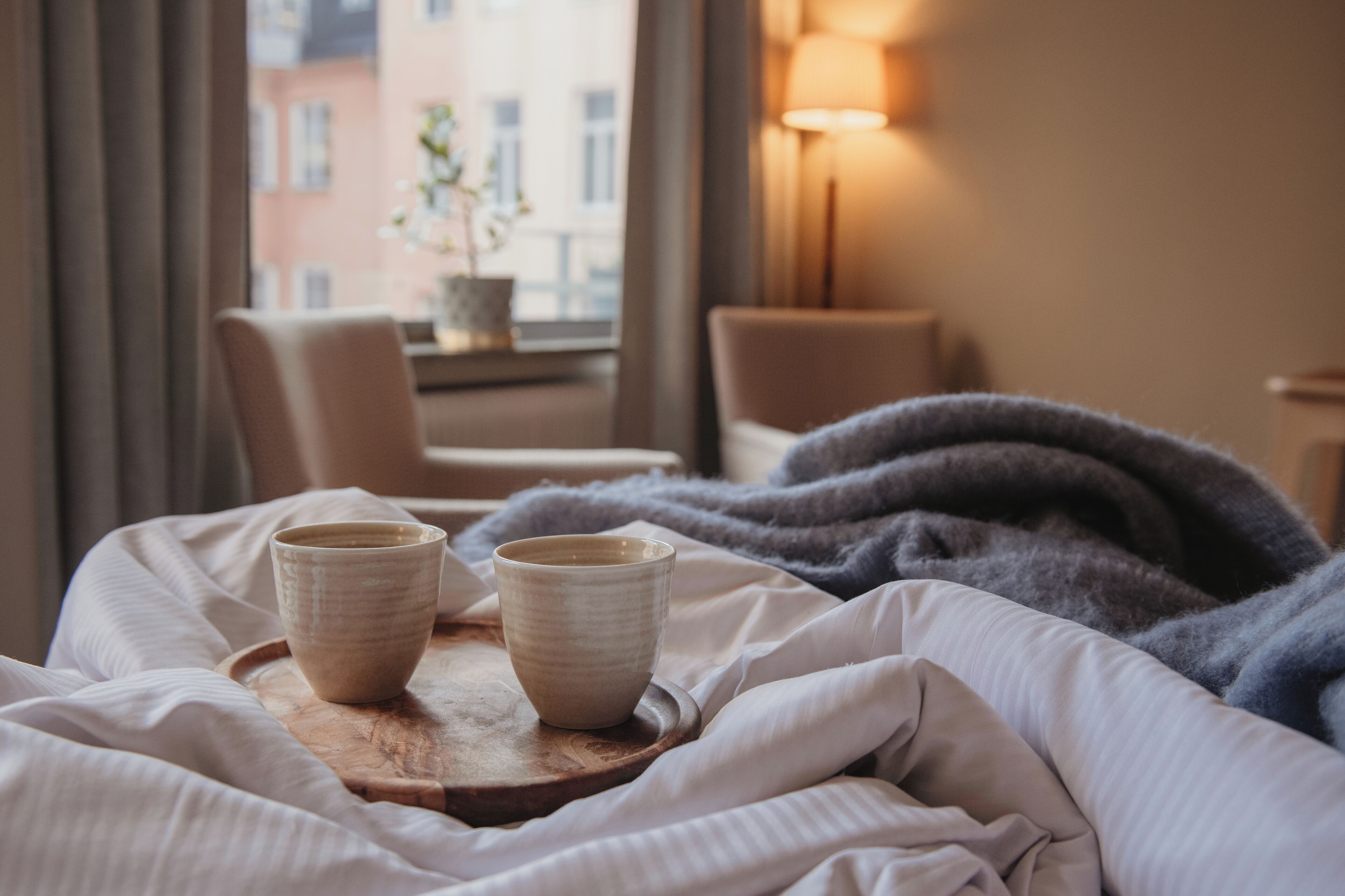 A hotel room with a bed, two cups on a tray, a chair, a lamp, and a view of buildings through a window.