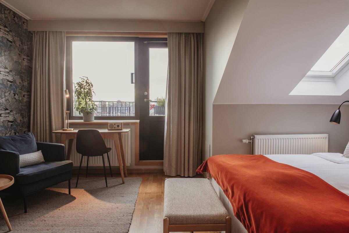 Un dormitorio con cama, escritorio, silla, banco y vistas al exterior.