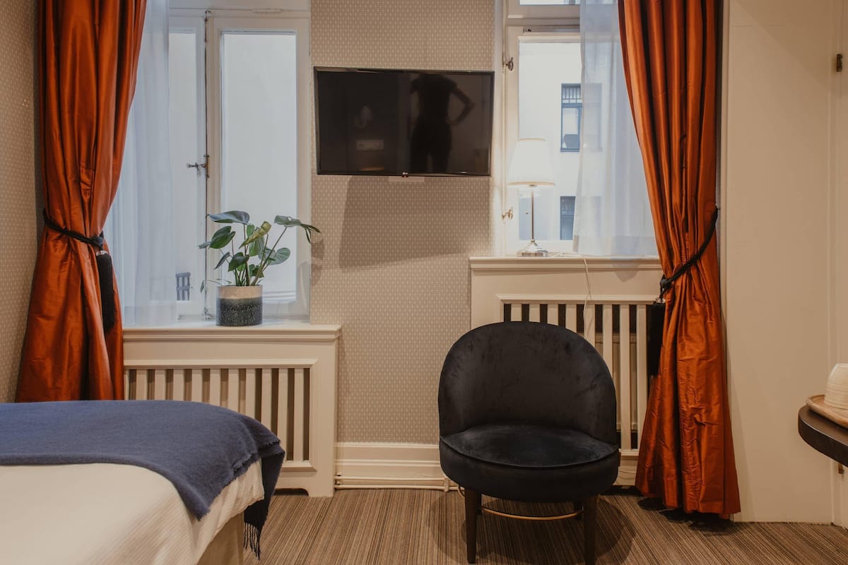 Una habitación de hotel con cama, una silla, un televisor, una ventana con cortinas y un radiador.