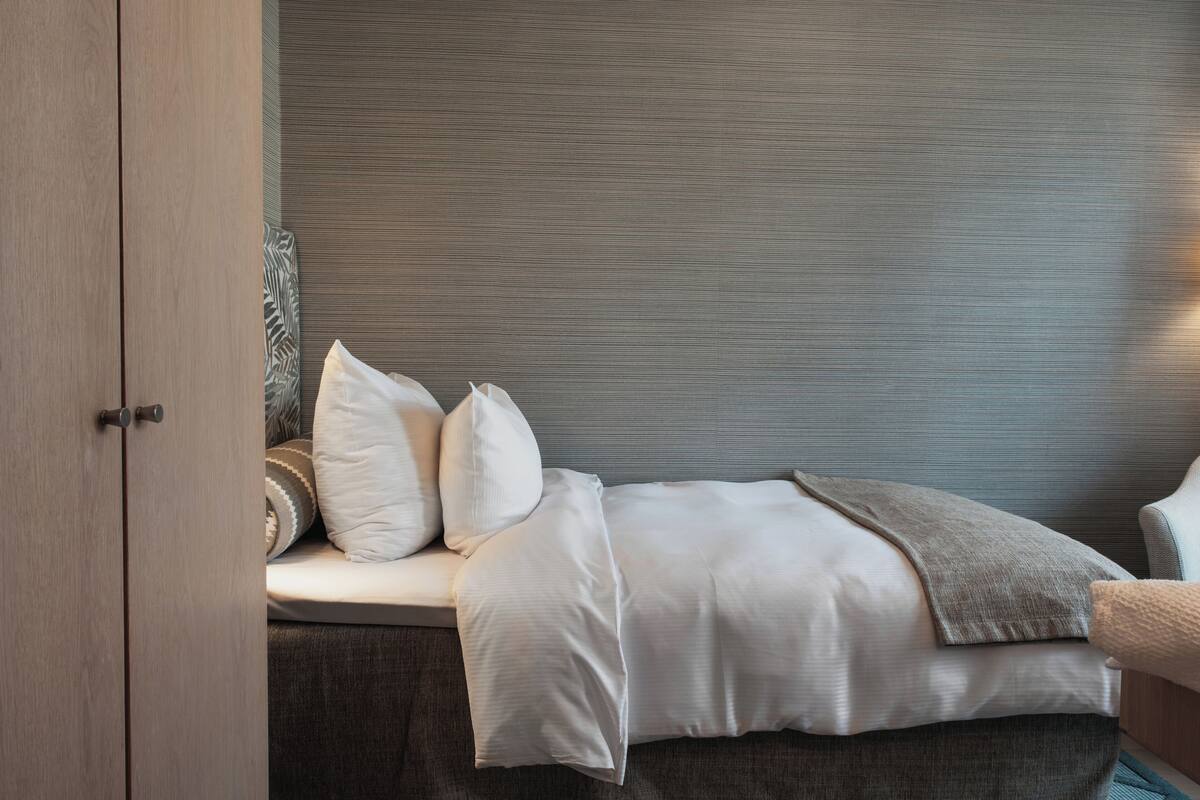 Una cama cuidadosamente tendida con ropa de cama blanca y un plaid gris, un banco tapizado en gris con un cojín estampado, y un armario de madera con un tirador.
