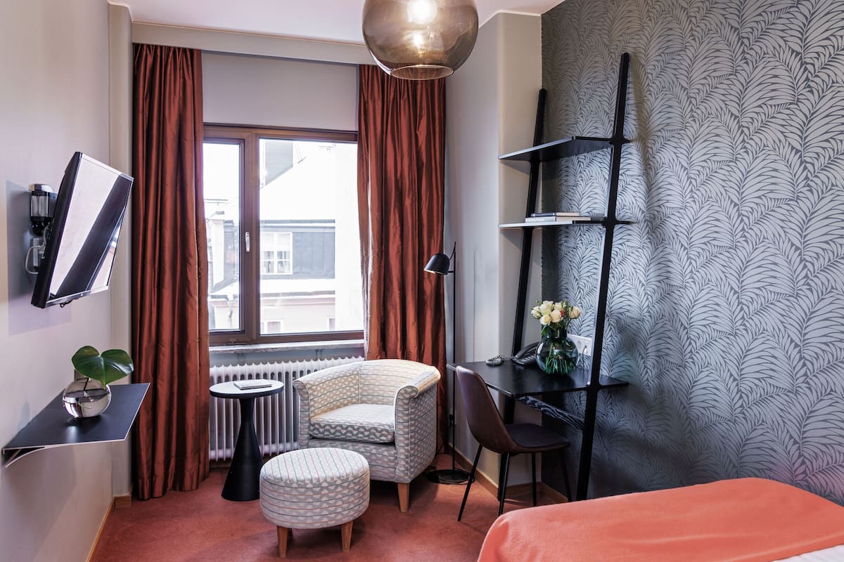 Habitación de hotel con cama, escritorio, silla, mesita, radiador, planta y ventana con cortinas.