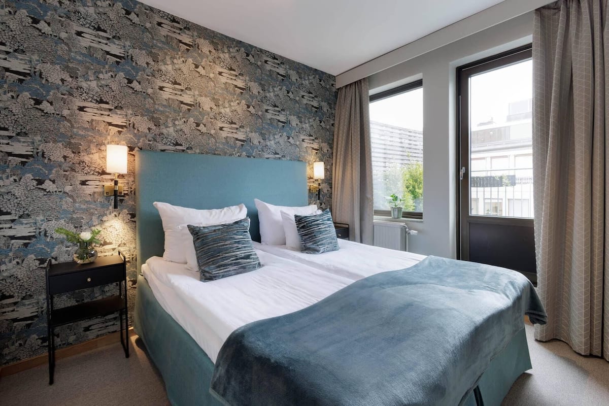 Habitación de hotel con pared de piedra, cama con ropa de cama blanca y azul, mesita de noche con lámpara y ventana con cortinas.