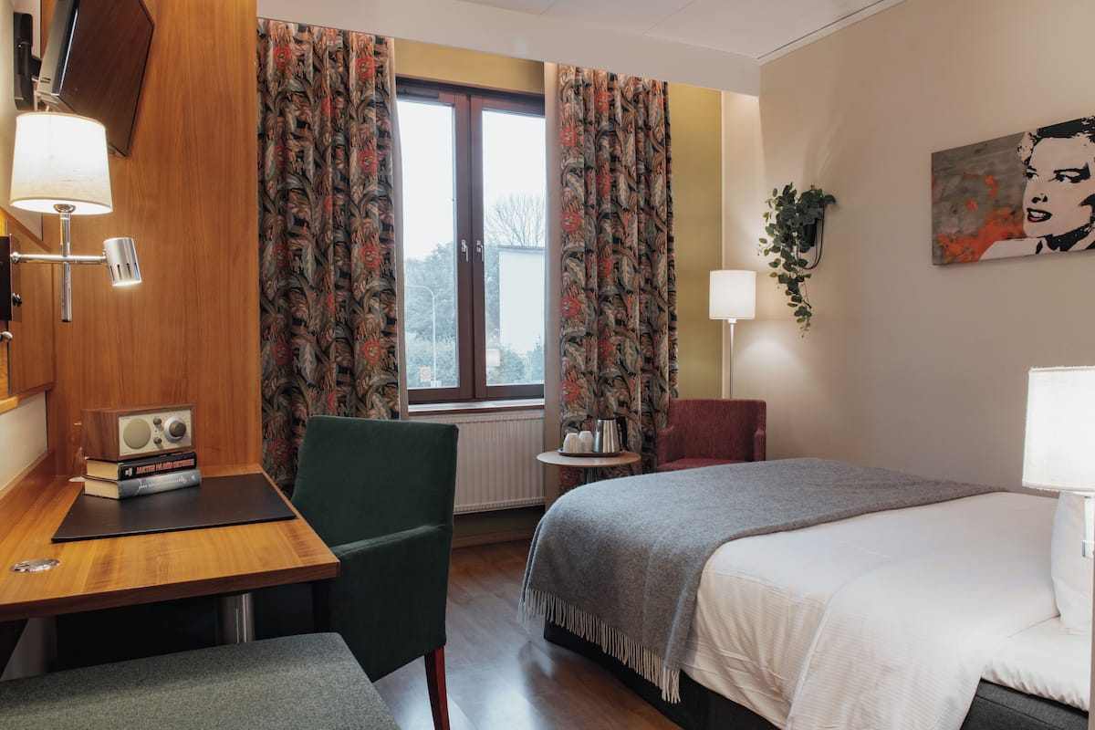Une chambre d’hôtel avec un lit, un bureau doté d’une lampe, une chaise, une fenêtre avec des rideaux et un tableau accroché au mur.
