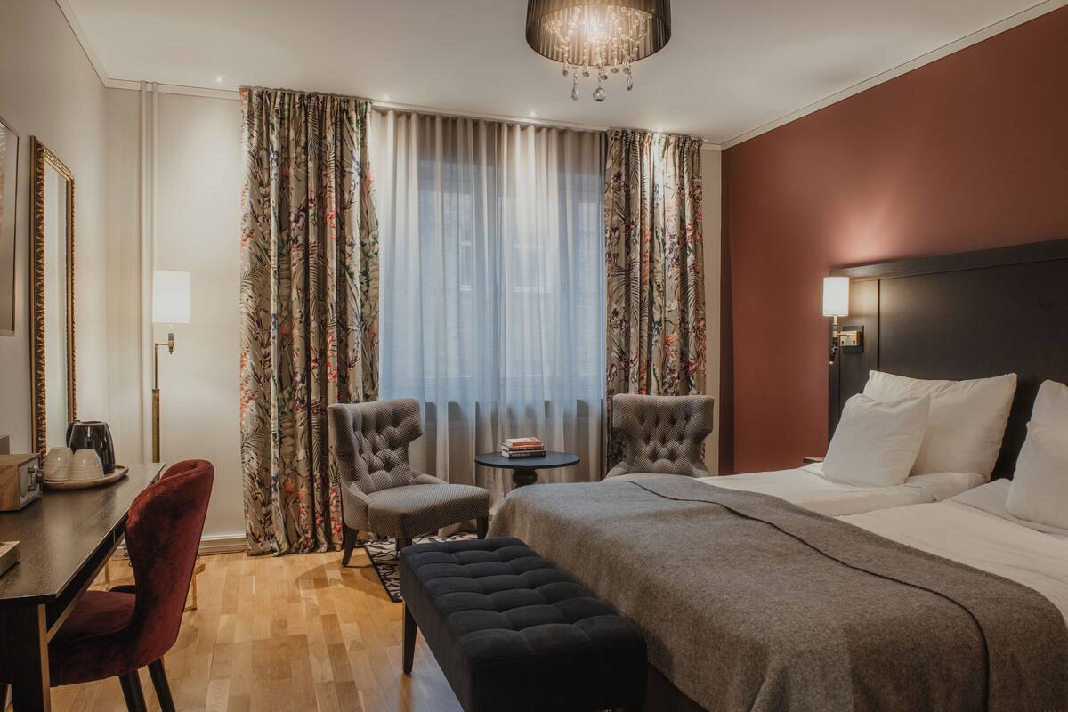 Une chambre d’hôtel avec un grand lit, deux fauteuils, une petite table et un bureau avec une lampe.