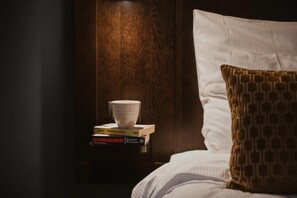Un lit bien fait, avec un oreiller blanc et un oreiller marron à motifs, une pile de livres et un bol blanc posé sur une table de chevet en bois.