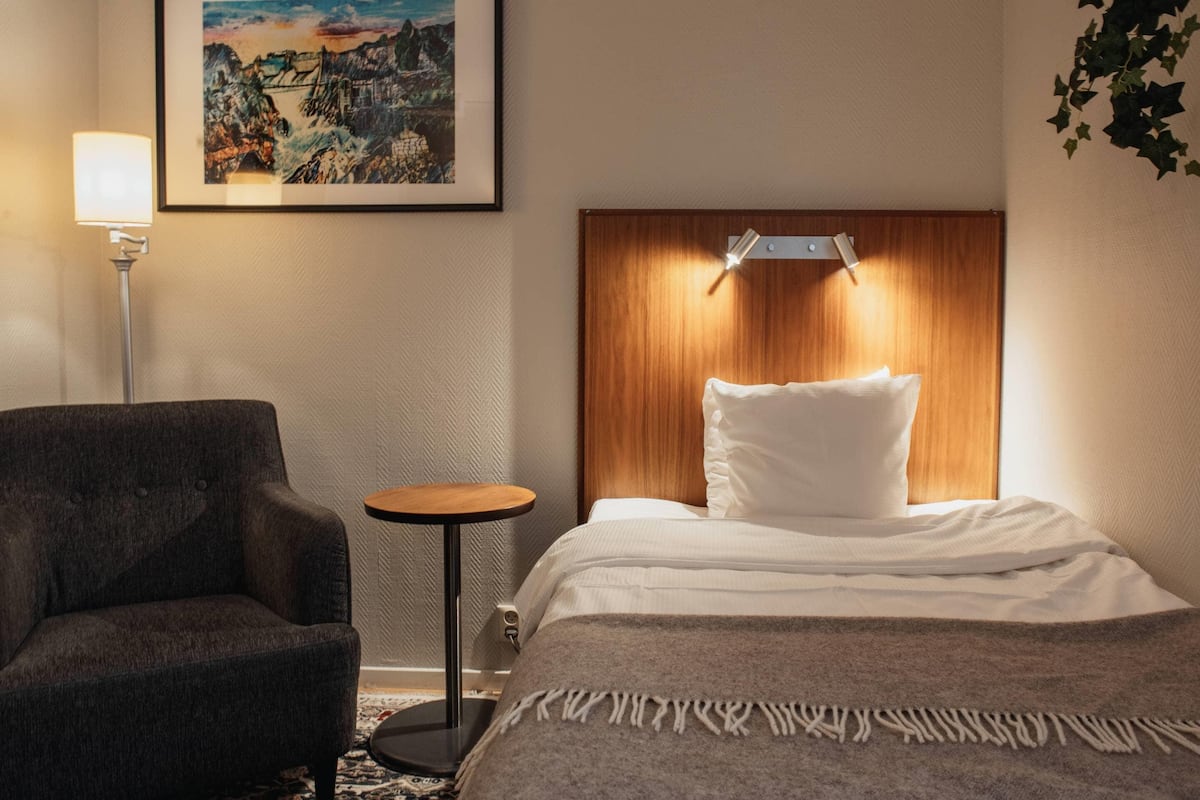 Une chambre d’hôtel avec un lit, une chaise, une petite table et une lampe fixée au mur.