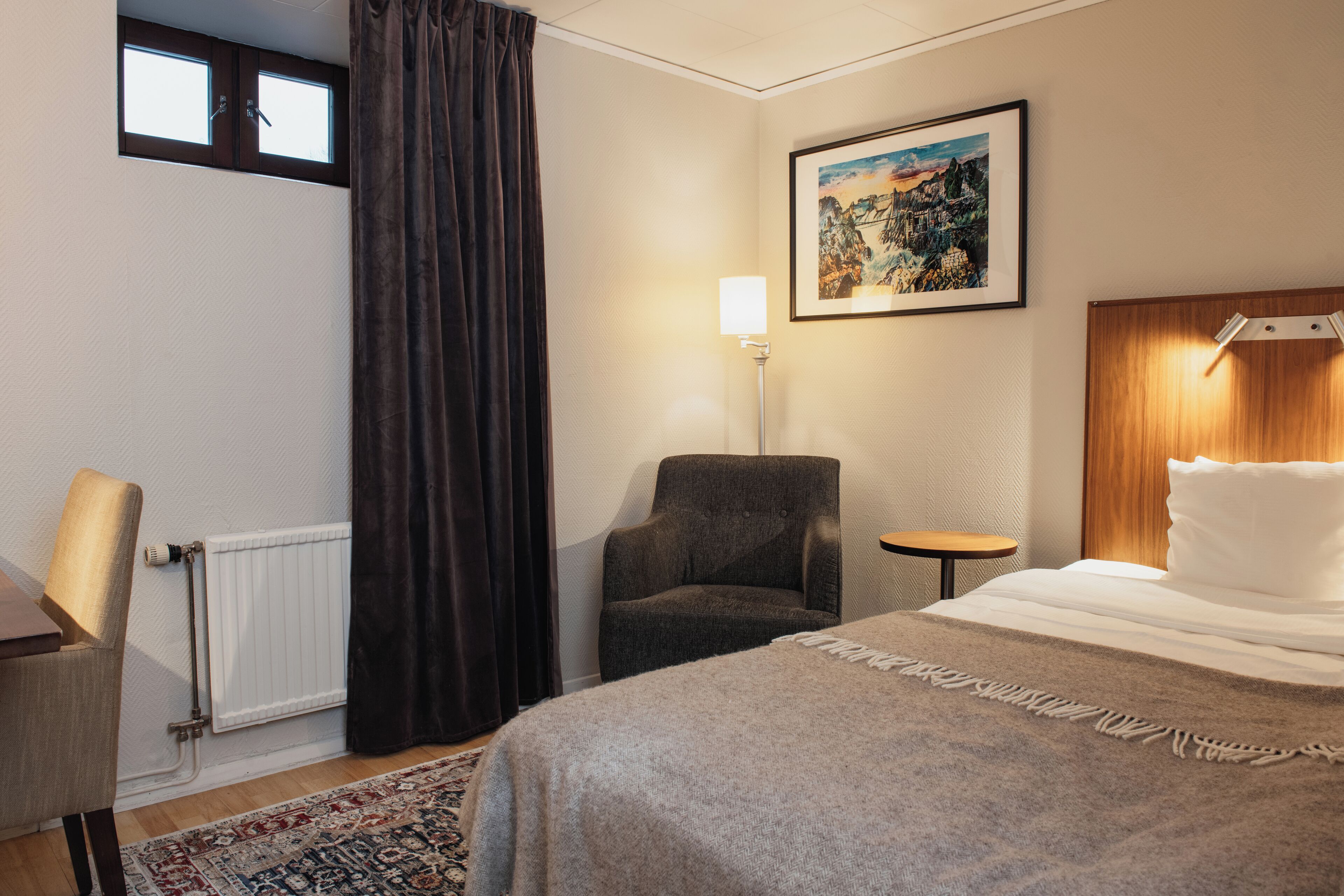Ett hotellrum med en säng, en stol, ett litet bord, en lampa, en tavla på väggen och ett fönster med gardiner.