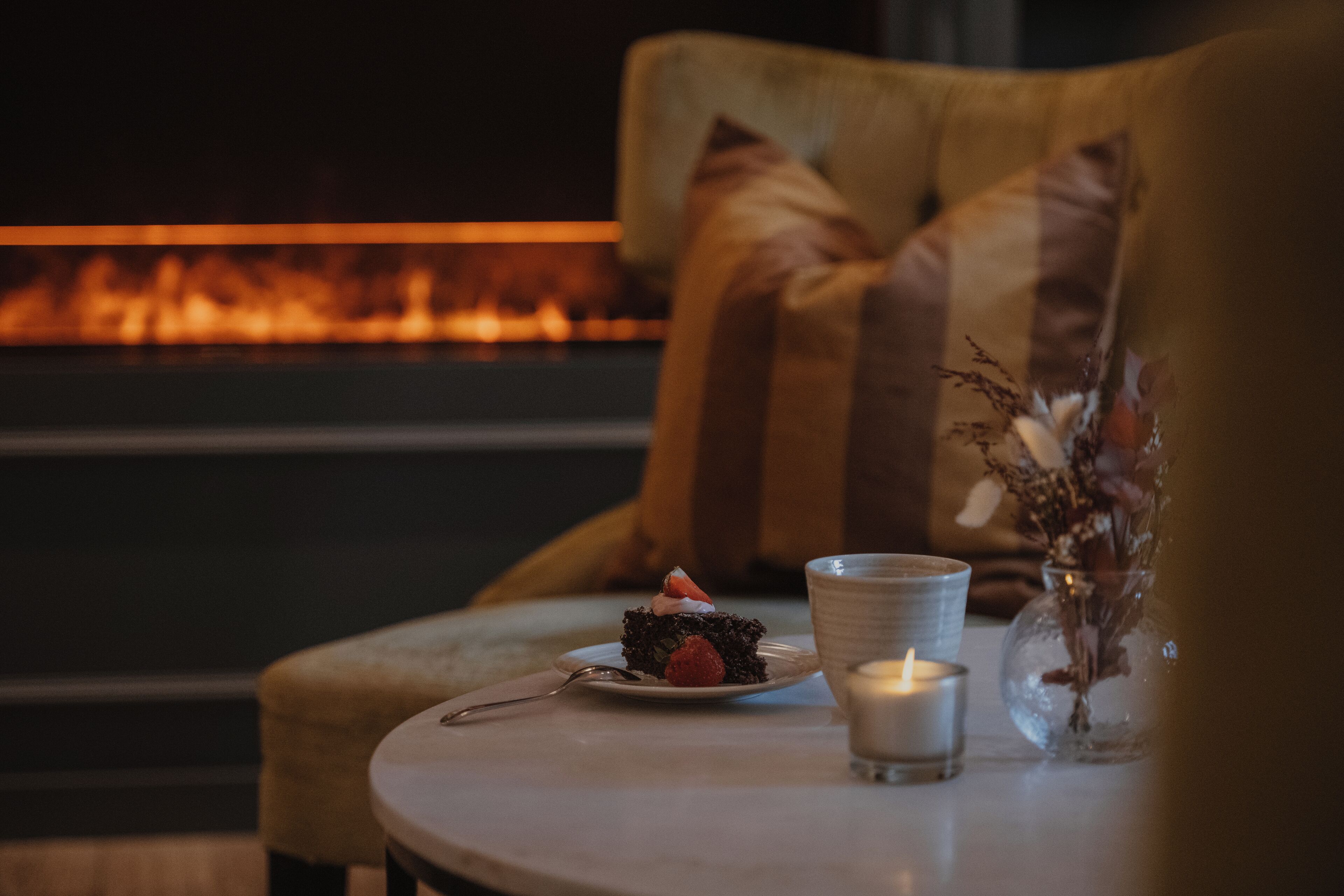 A cozy seating area with a cushioned chair, a small table with a plate of food, a candle, and a vase with dried flowers.