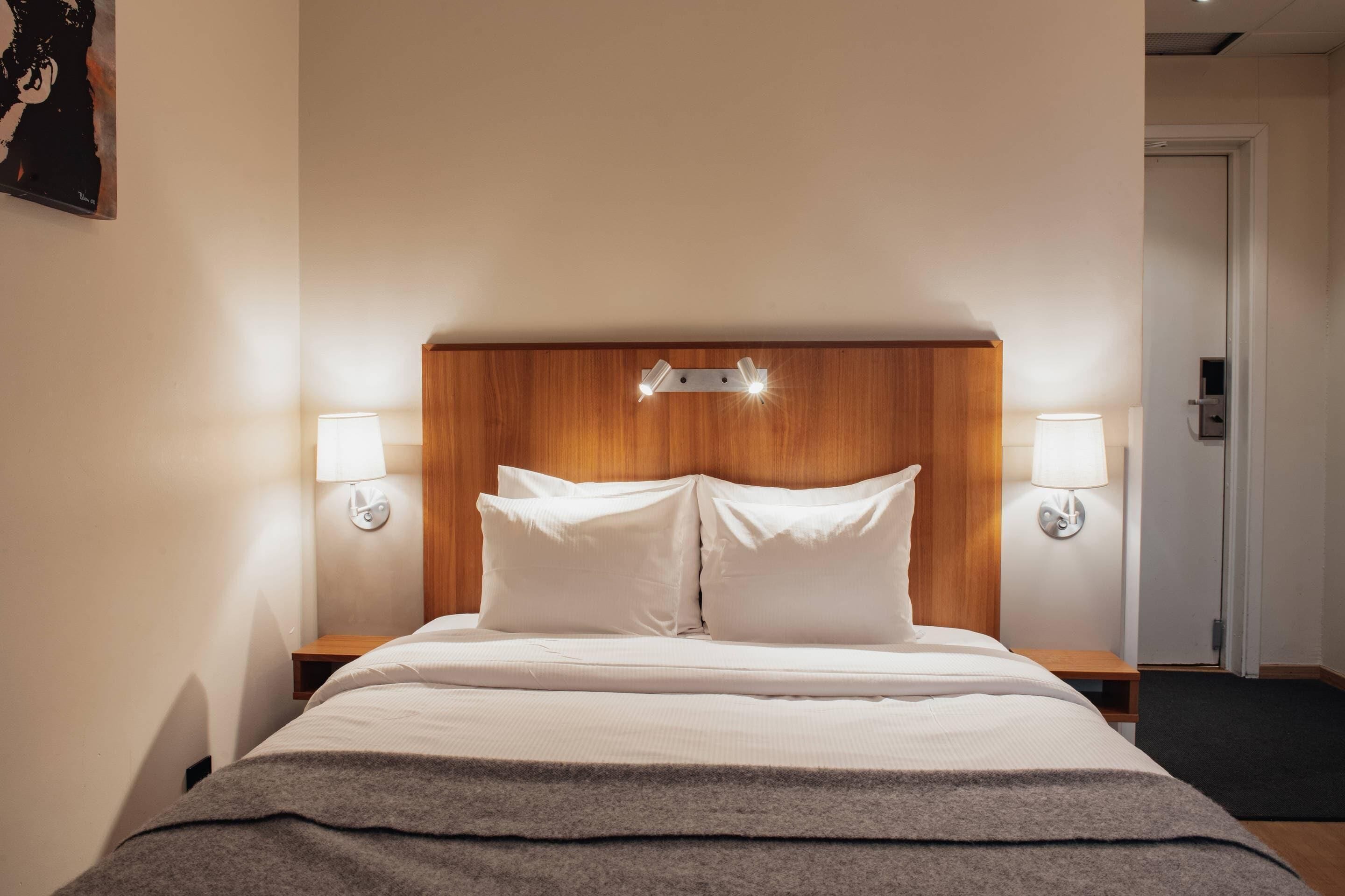 Una cama bien hecha con sábanas blancas y una manta gris, con dos mesitas de noche a cada lado y lámparas fijadas a la pared.