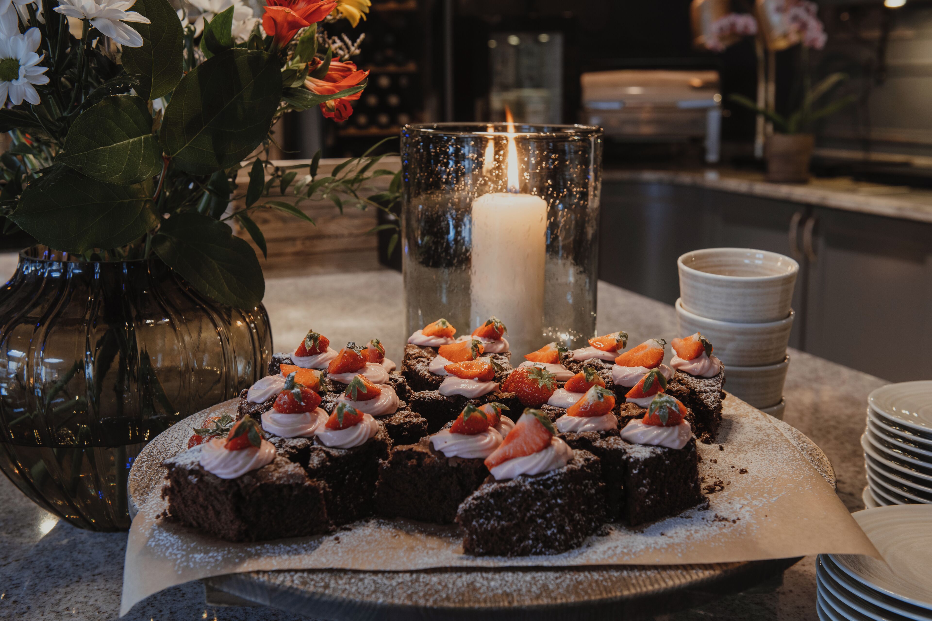 A dessert table with a lit candle, a vase with flowers, and a stack of plates.