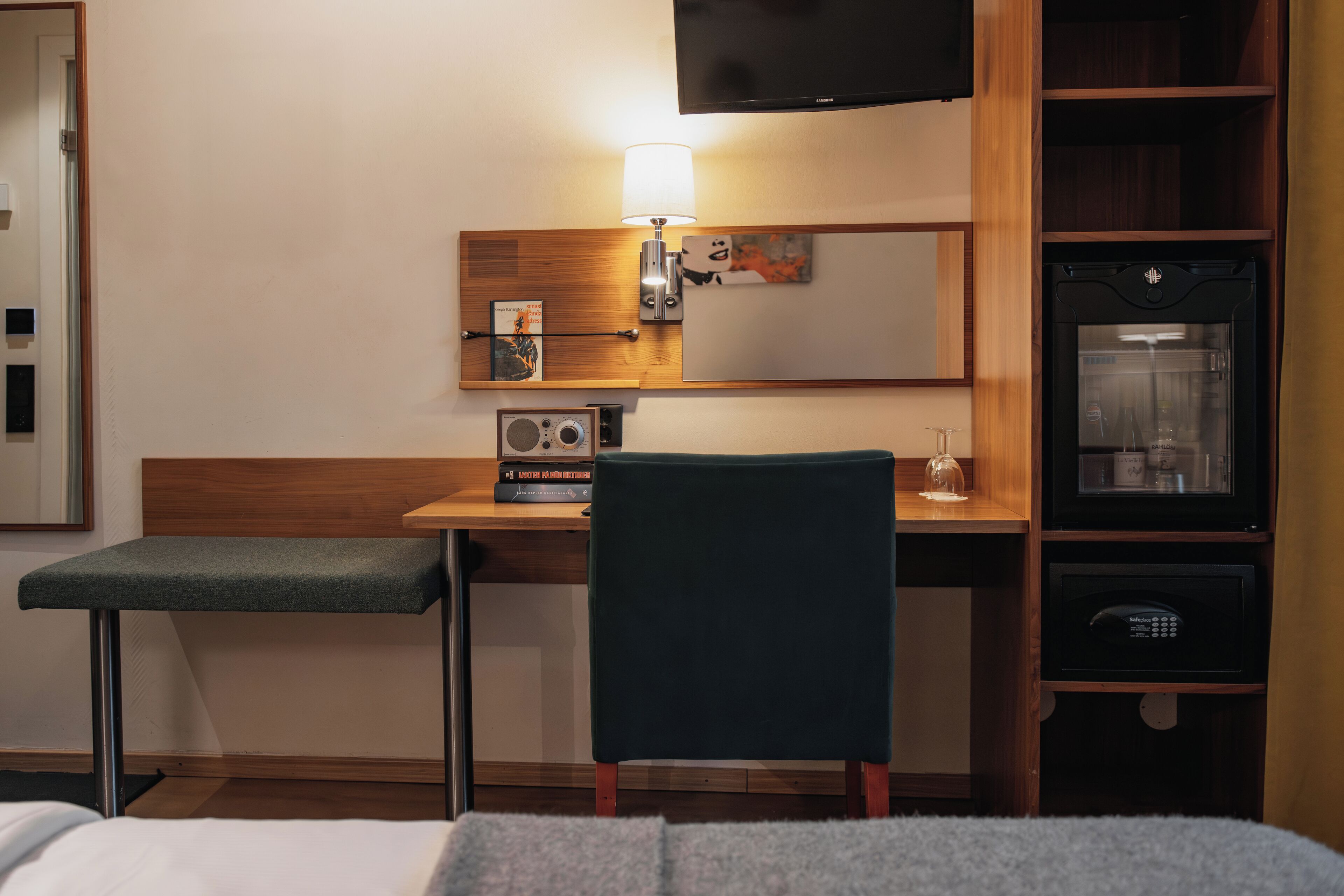 Una camera d'albergo con un letto, una scrivania con sedia, una televisione, un minibar e una lampada a parete.
