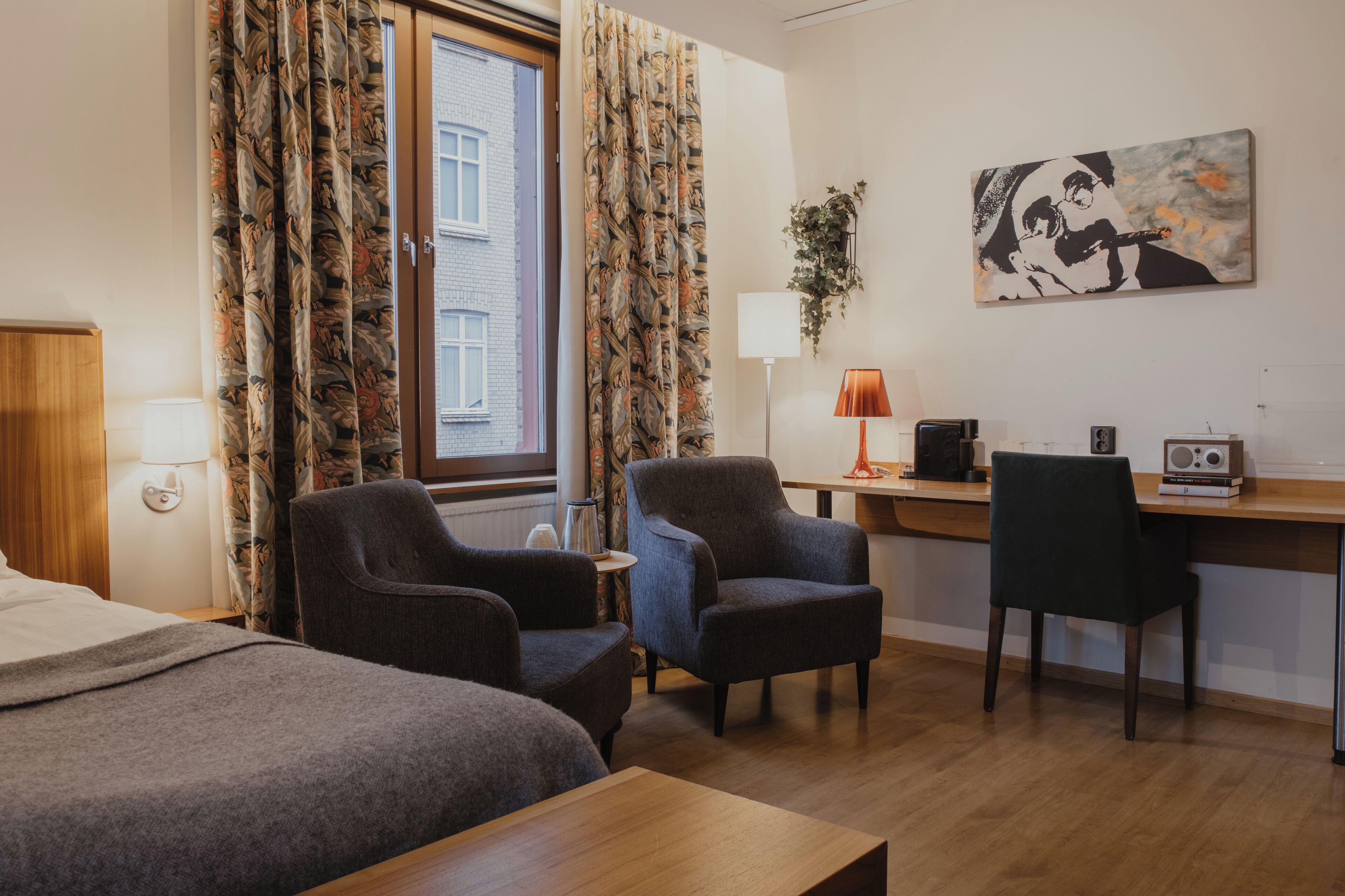Ett hotellrum med två sängar, ett skrivbord, en stol, en lampa och en tavla på väggen.