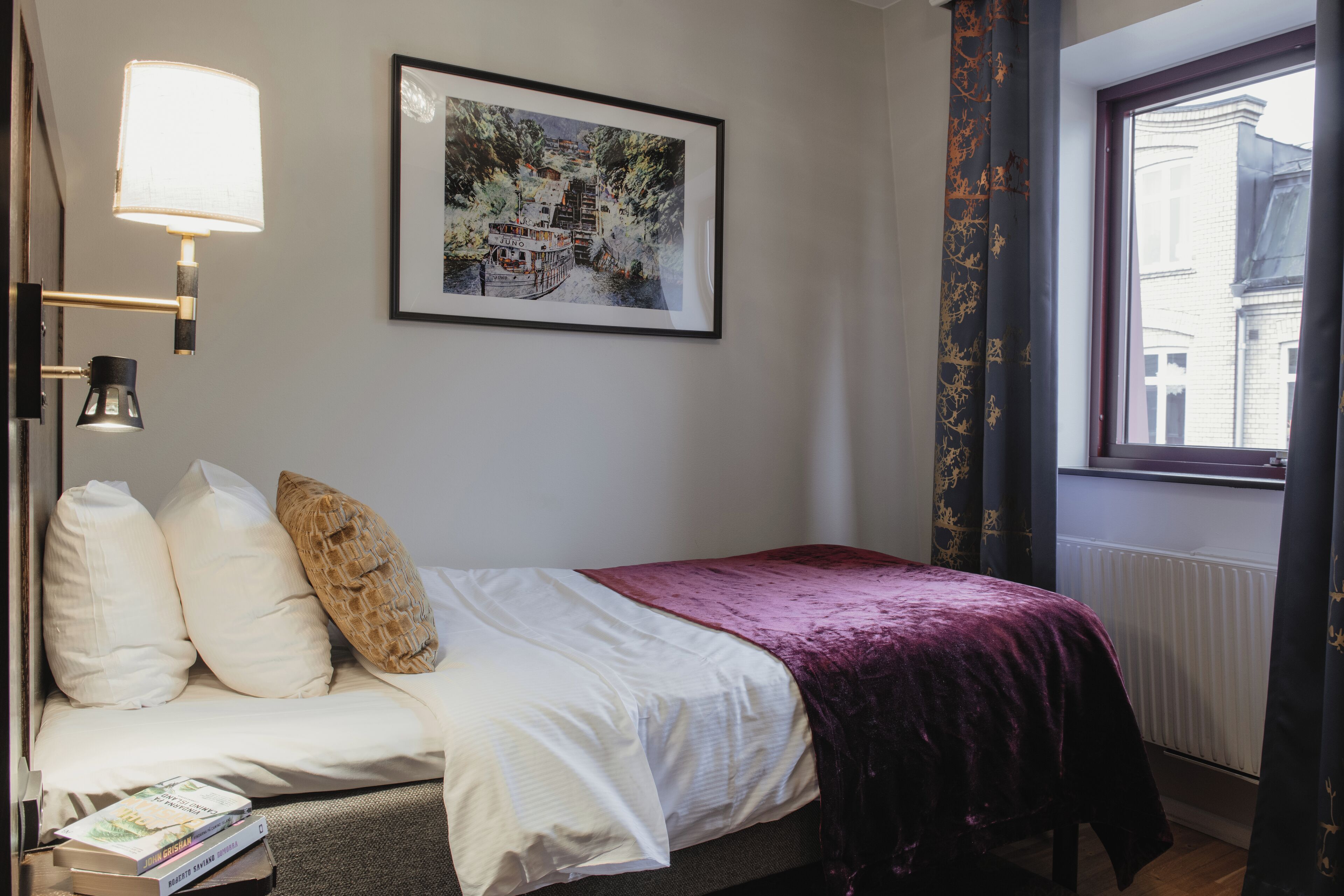 A neatly made bed with a white and purple bedspread, a framed picture on the wall, and a window with curtains.