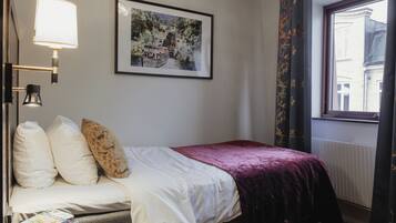 A neatly made bed with a white and purple bedspread, a framed picture on the wall, and a window with curtains.