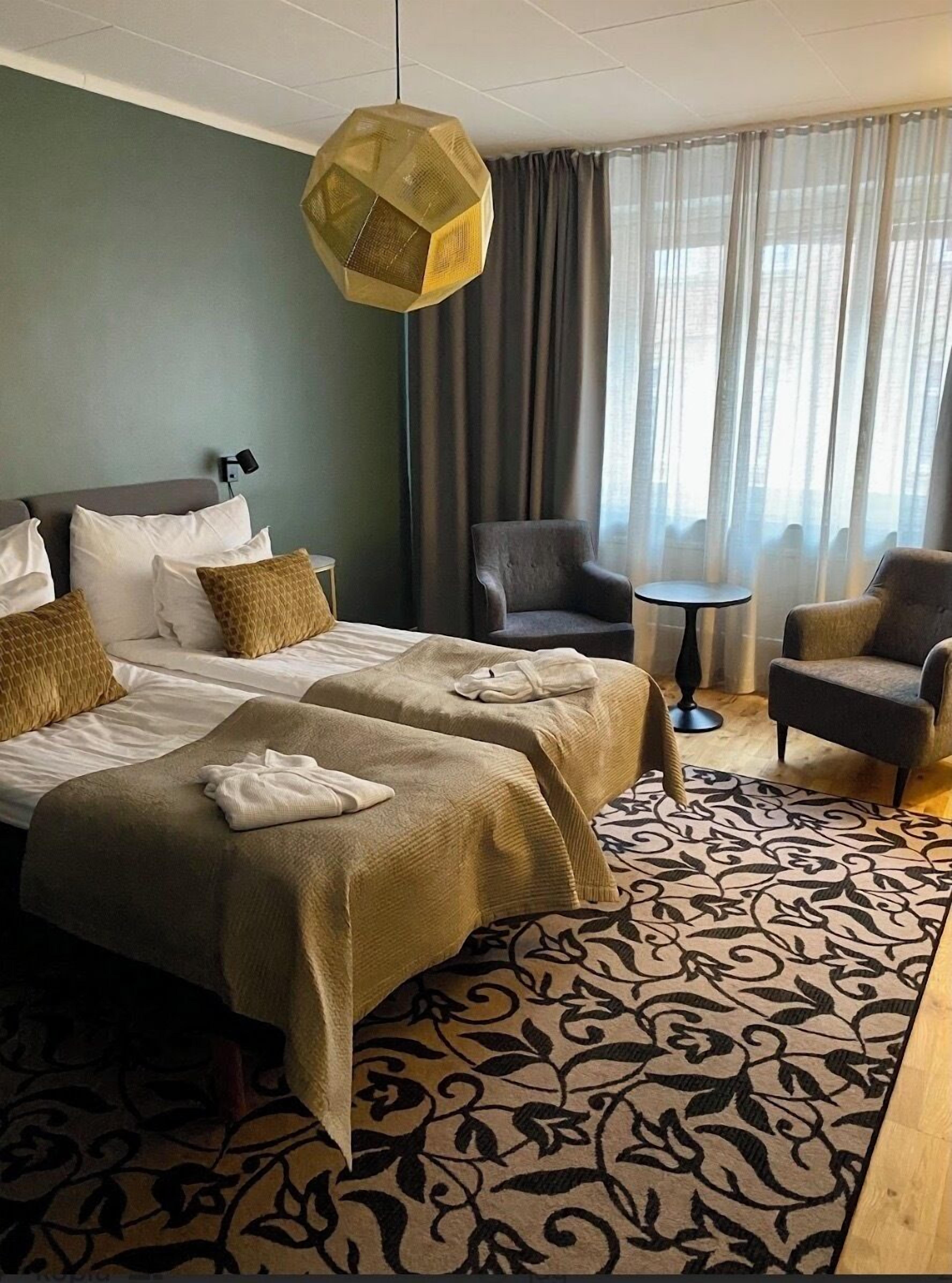 A hotel room with two beds, a chair, a small table, and a patterned rug.