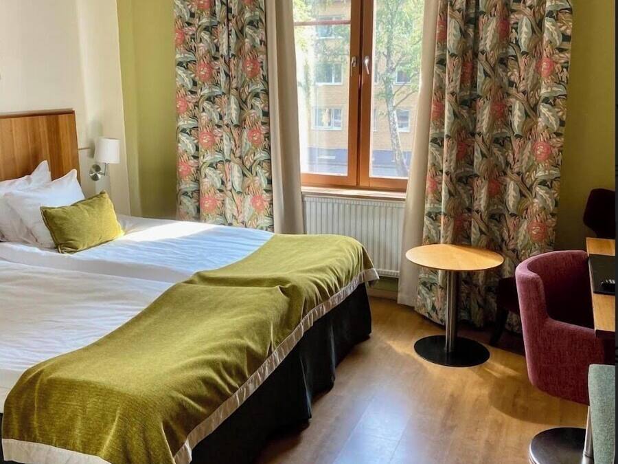 Ett hotellrum med en säng, ett litet runt bord, en stol och gardiner med ett blommönster.