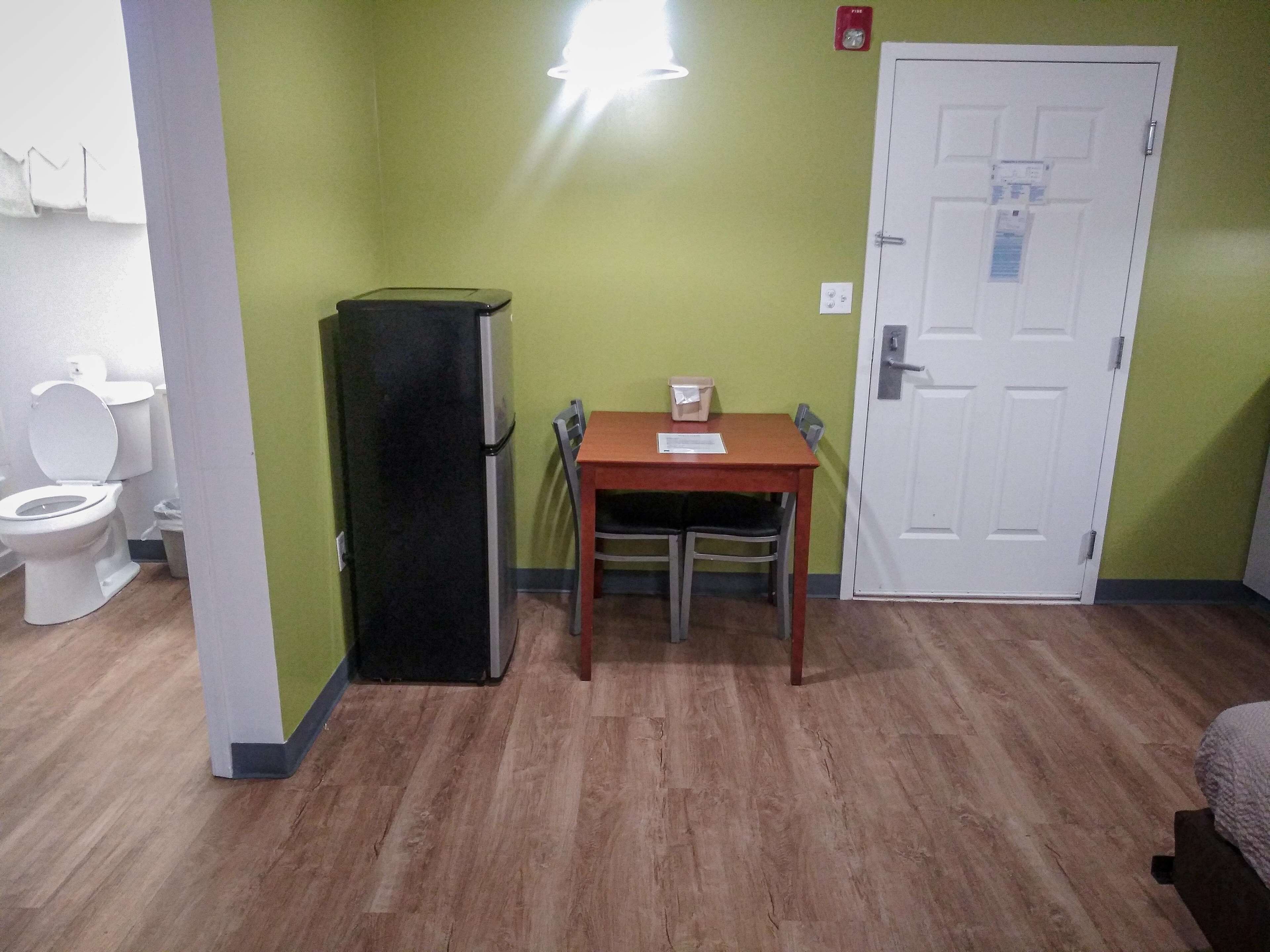 Una habitación pequeña con piso de madera, una puerta blanca, un refrigerador negro, una mesa de madera con dos sillas y un inodoro blanco.