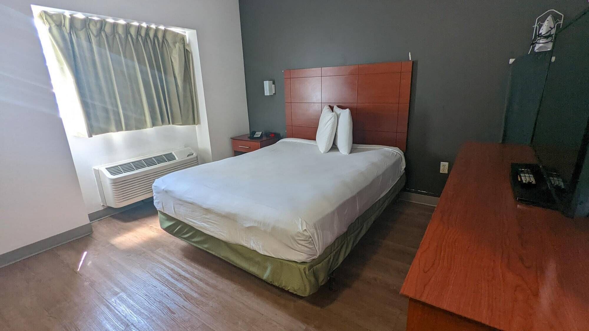 A hotel room with a bed, a wooden headboard, a bedside table, a nightstand, a telephone, and a window with curtains.