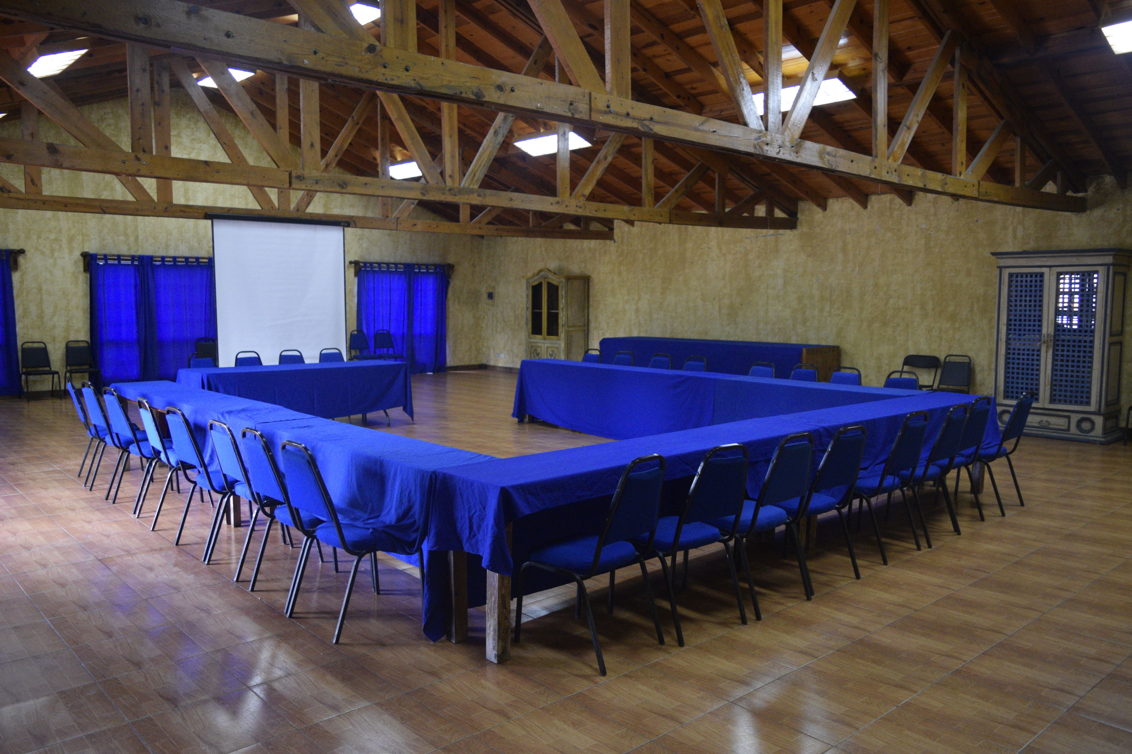 A large hall with a long table and chairs, wooden beams on the ceiling, and a projector screen.