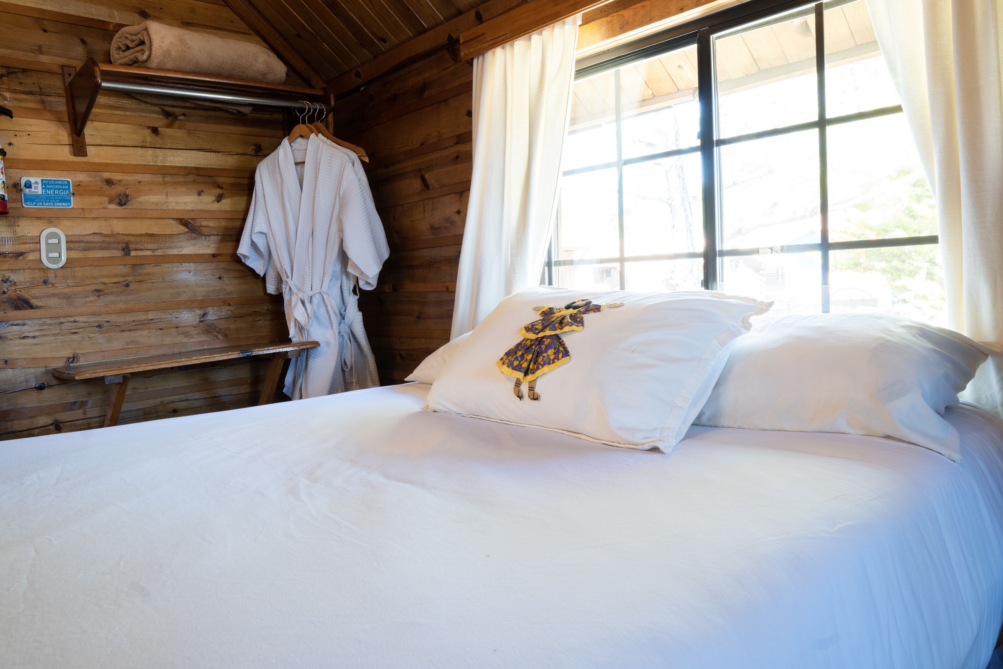 A wooden cabin room with a bed, a window with curtains, a towel rack, and a sign on the wall.