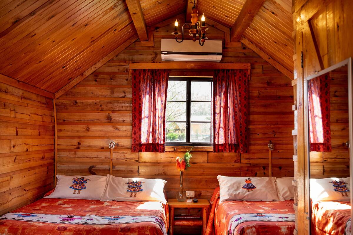 Une chambre de style cabane en bois, avec deux lits, une fenêtre avec des rideaux, un appareil de climatisation et un lustre.