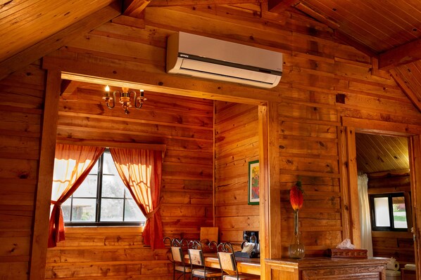 L’intérieur d’un chalet en bois comprend un coin repas, un lustre, un appareil de climatisation et une fenêtre avec des rideaux.