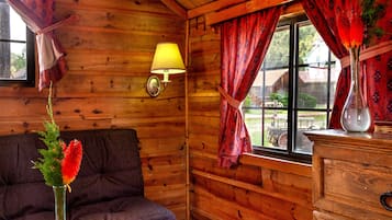 A wooden cabin interior with a sofa, a lamp, a vase with flowers, and a window with curtains.