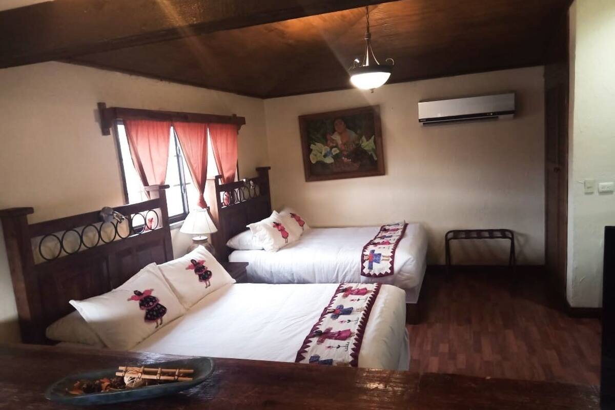 Une chambre avec deux lits, des têtes de lit en bois, un plancher en bois, un tableau au mur et une fenêtre avec des rideaux.