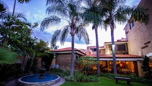 A courtyard with a fountain, palm trees, and a building with a red-tiled roof. - La Mansiรณn del Sol (Zapopan)