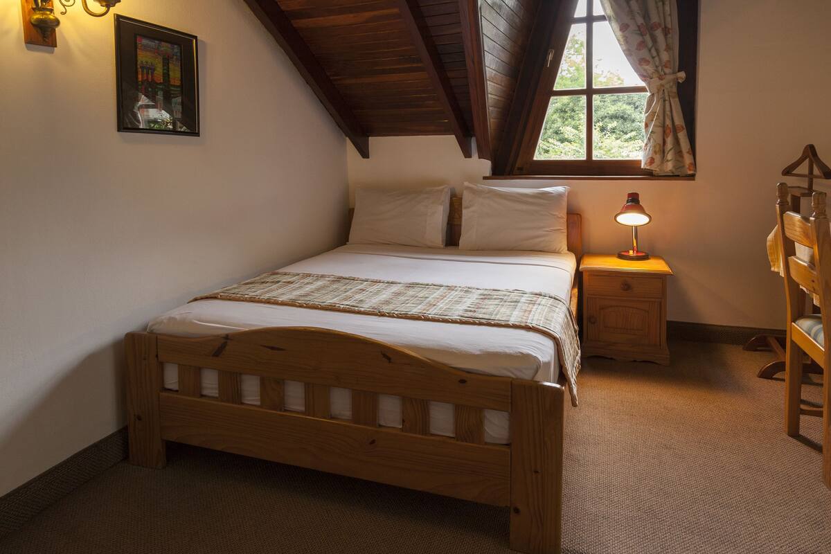A bedroom with a wooden bed, a nightstand with a lamp, a dresser, and a window with curtains.