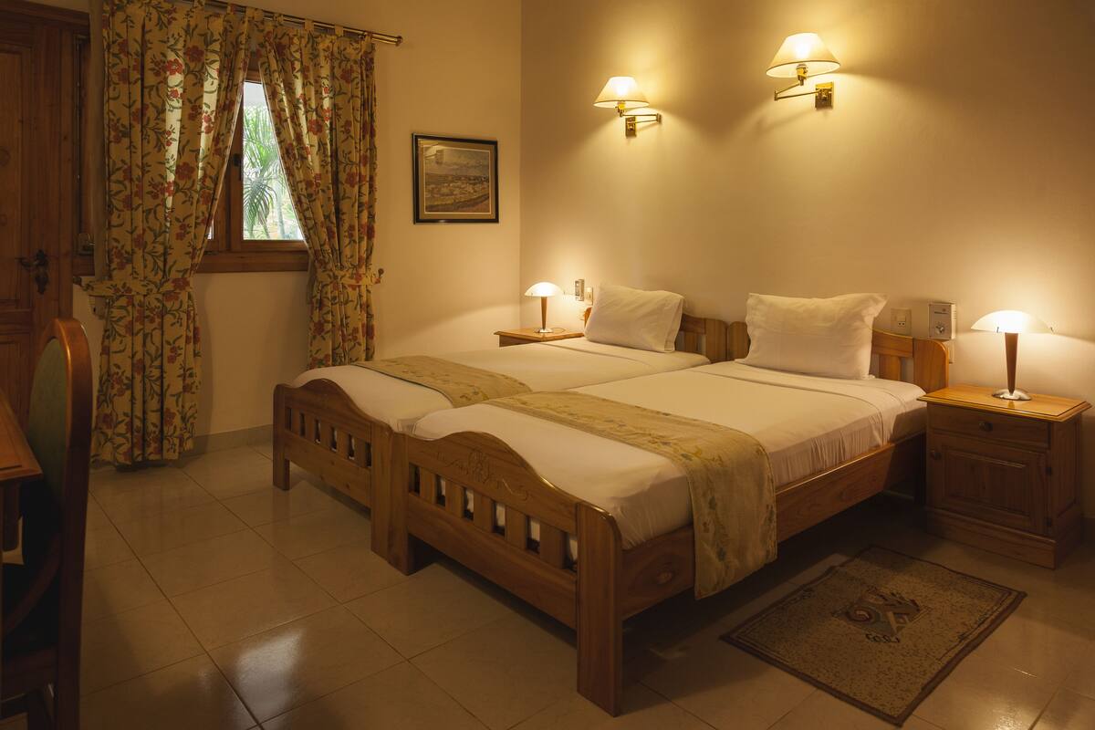 A double bed with two bedside tables, a nightstand, a wooden headboard, a window with curtains, a wall-mounted lamp, and a framed picture.
