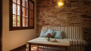 Una habitación acogedora con pared de piedra, un sofá a rayas, una mesa de madera con un jarrón de flores y una ventana con contraventanas de madera.