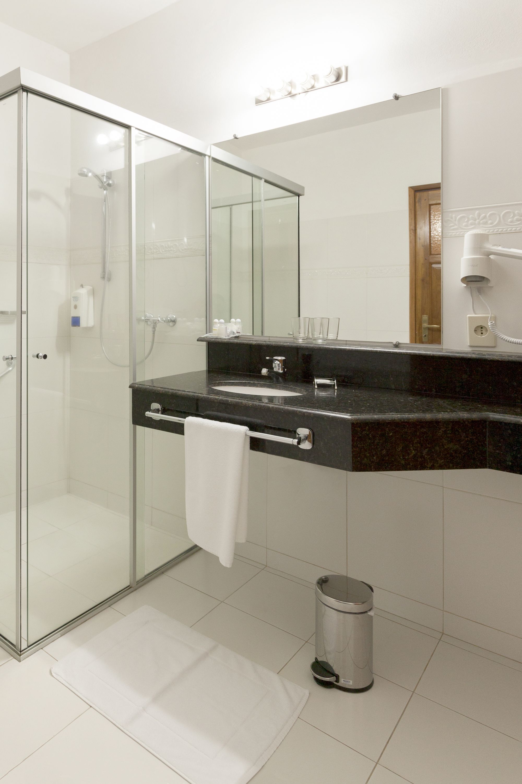 A modern bathroom with a glass shower enclosure, a black countertop sink, a large mirror, and a hairdryer on the wall.