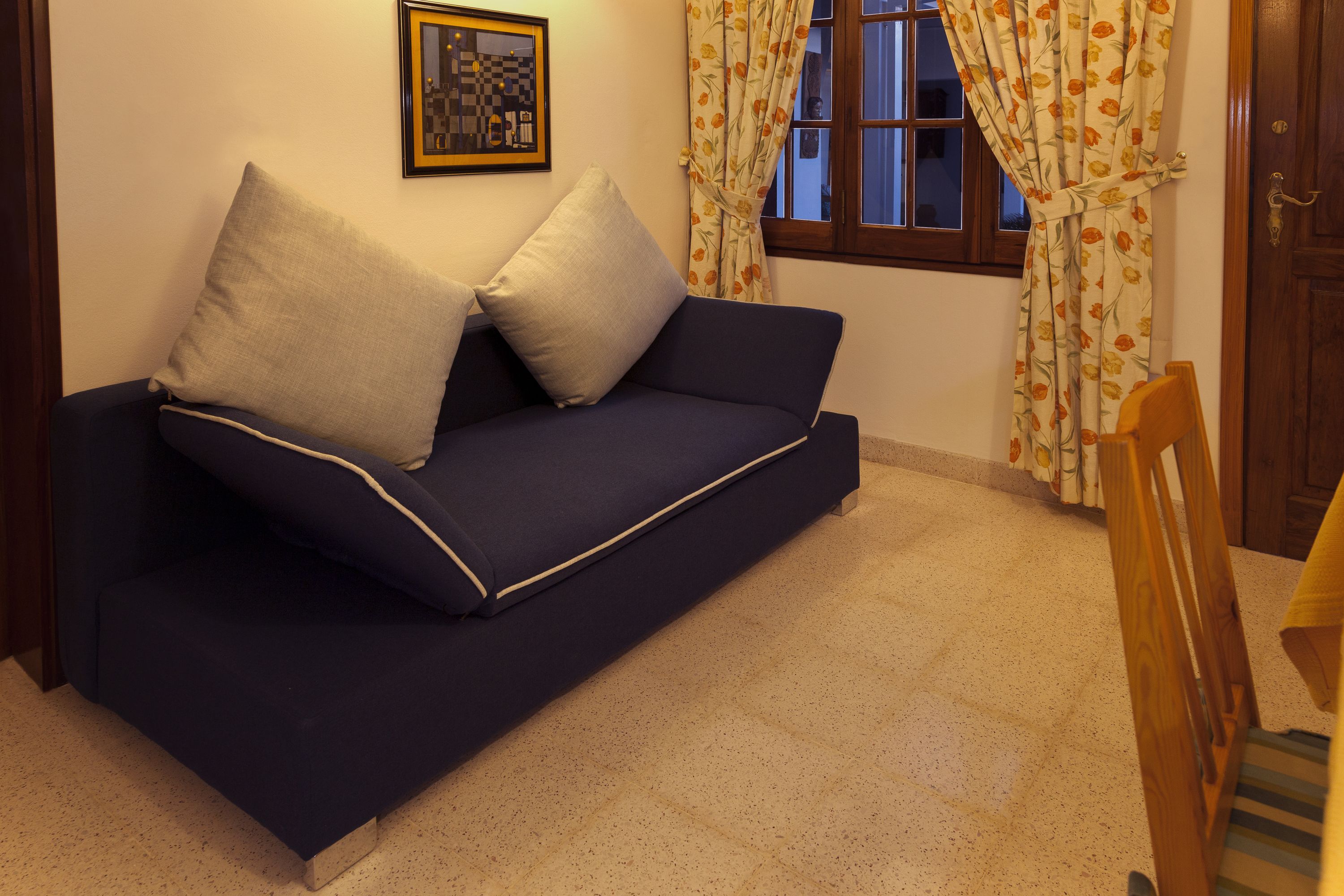 Un divano blu scuro con due cuscini, un quadro incorniciato al muro, una finestra con tende e una sedia di legno.