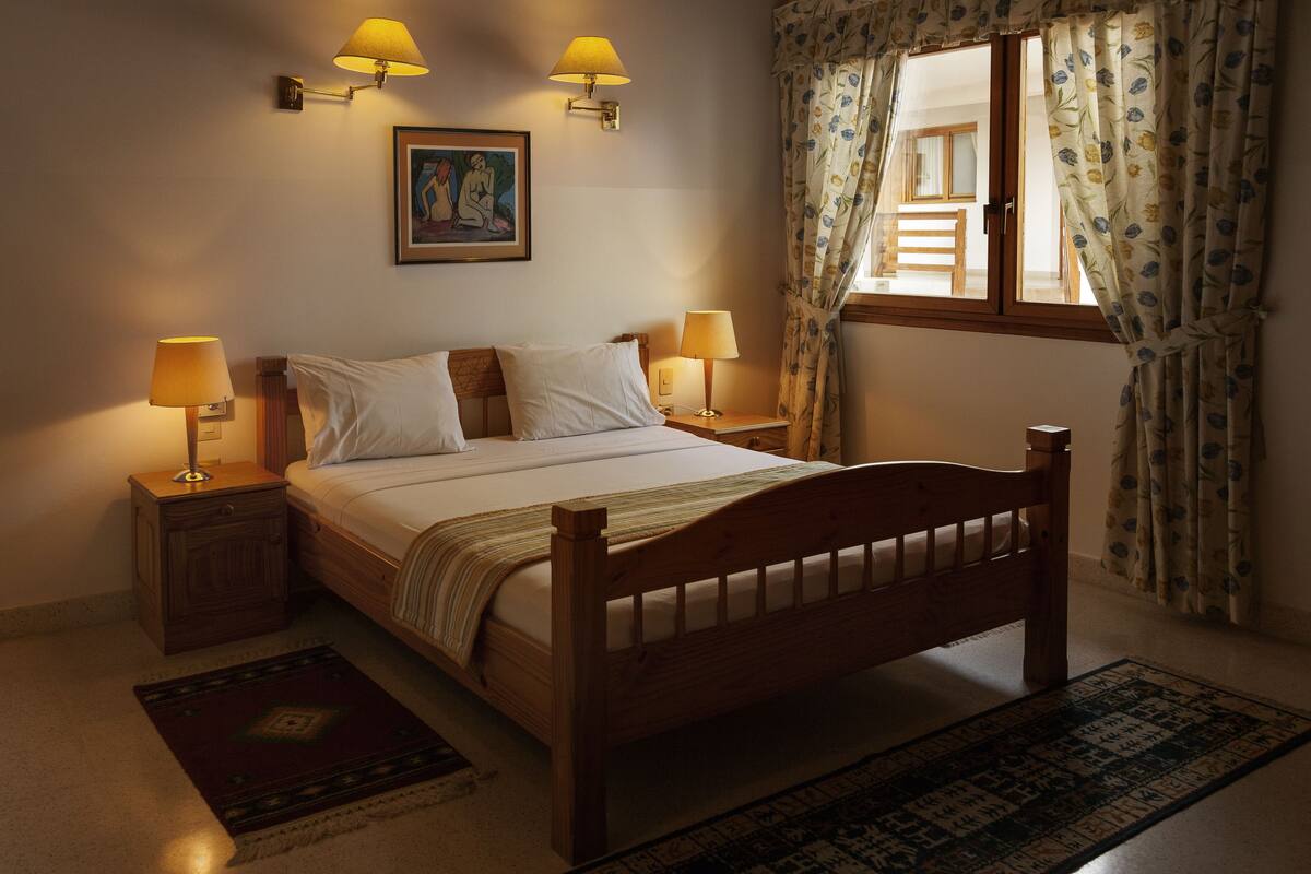 A bedroom with a wooden bed, two bedside lamps, a nightstand, a patterned rug, and a window with floral curtains.