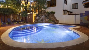 Una piscina con cascada de piedra, rodeada de sillones de descanso y una valla de madera.