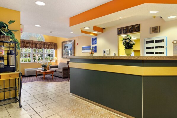 A reception area with a counter, framed pictures, a potted plant, and a window with a view of greenery. - Hometown Inn & Suites Jacksonville - Butler Blvd./Southpoint (Jacksonville)