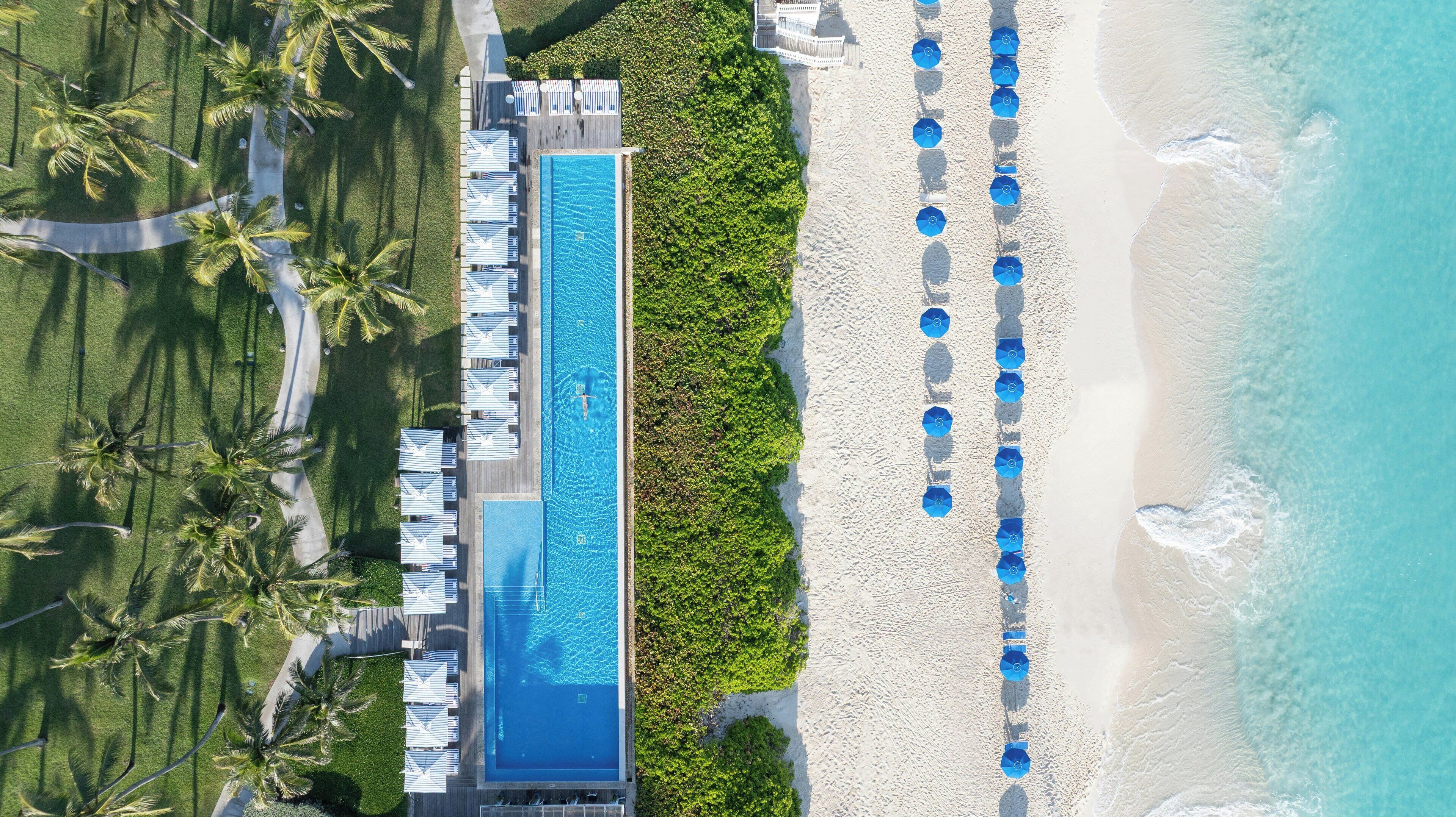 An aerial view of a resort with a long swimming pool, a beach with umbrellas, and palm trees.