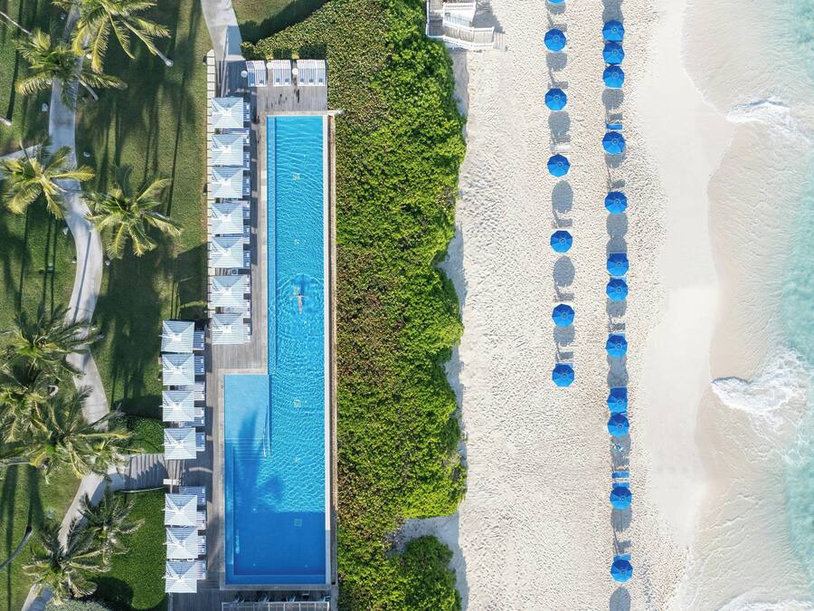 An aerial view of a resort with a long swimming pool, a beach with umbrellas, and palm trees.