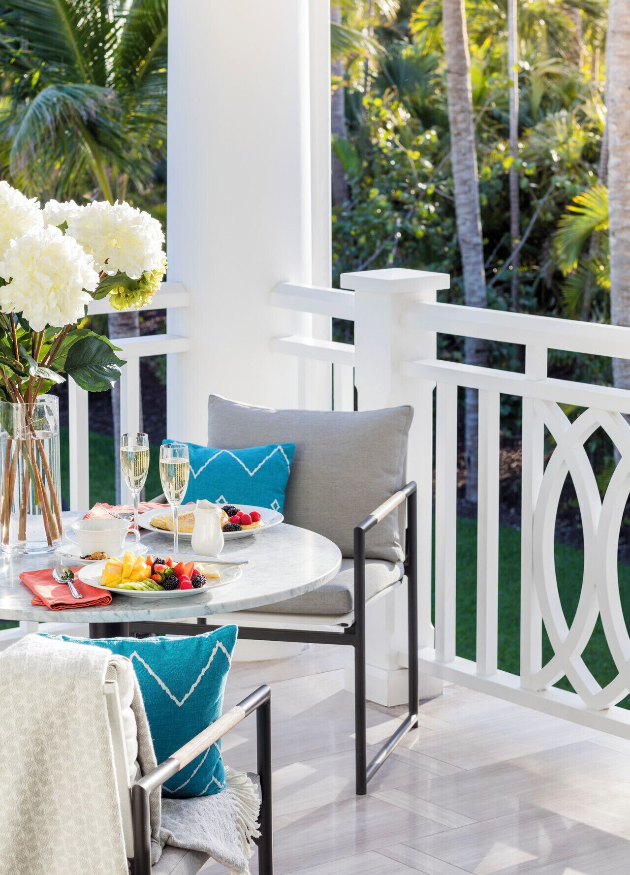 A patio with a round table set for two, featuring a white tablecloth, two glasses of white wine, a plate of fruit, and a pitcher of water. There are two chairs with cushions, a vase of white flowers, and a white railing in the background.