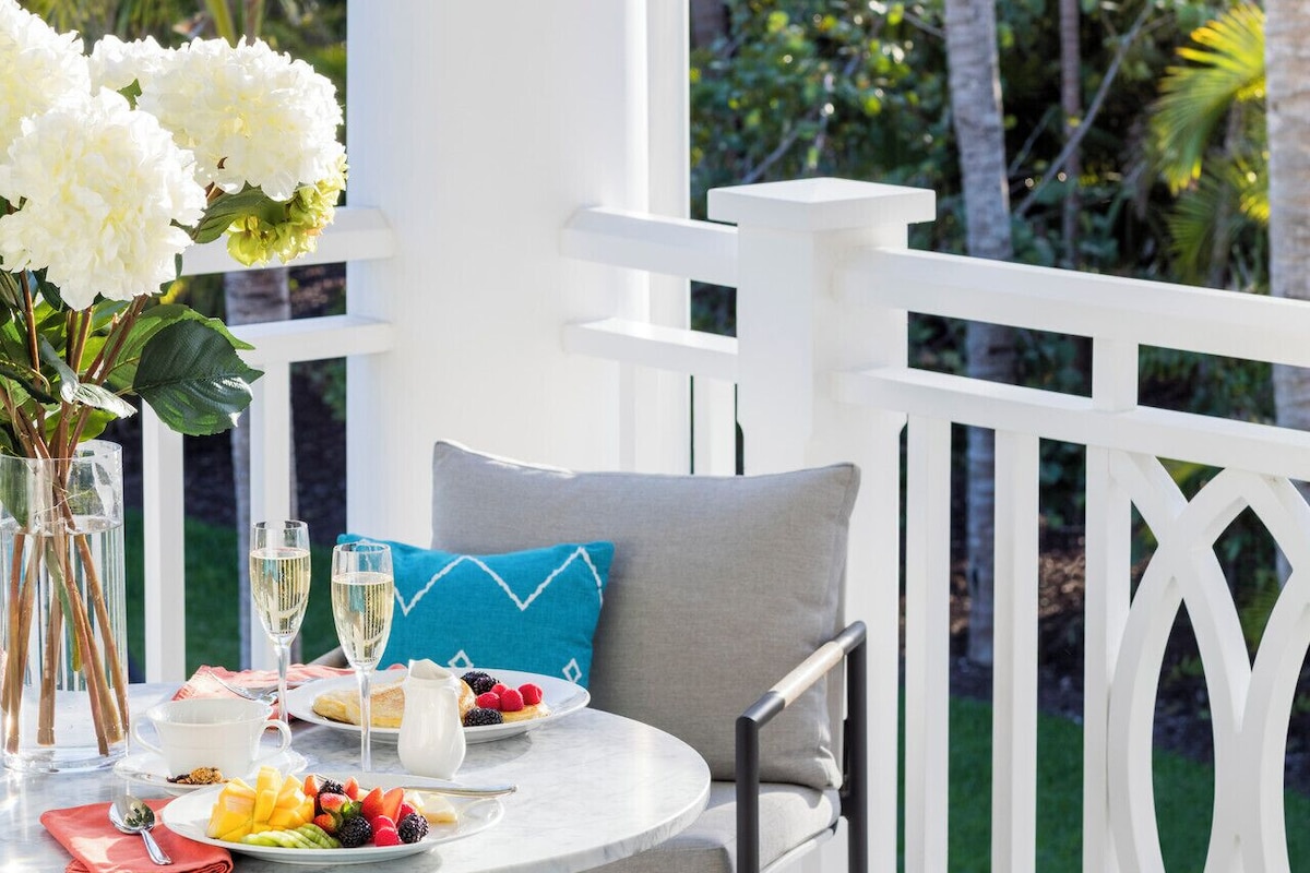 A patio with a round table set for two, featuring a white tablecloth, two glasses of white wine, a plate of fruit, and a pitcher of water. There are two chairs with cushions, a vase of white flowers, and a white railing in the background.
